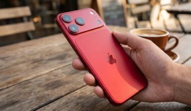 A hand holding a red smartphone with three rear cameras and an Apple logo, on a wooden table next to a cup of coffee.