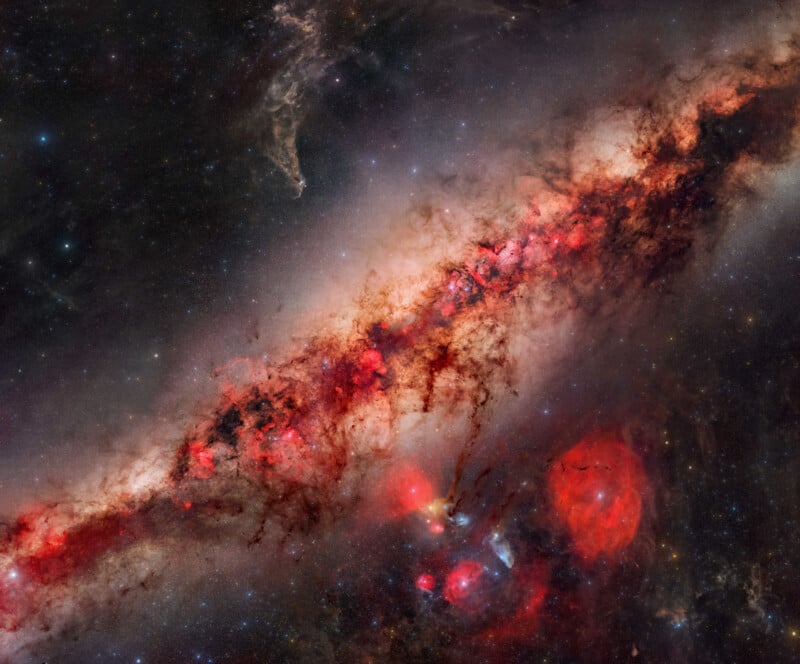 A colorful galaxy with a bright, reddish central band filled with glowing gas and dark dust lanes, surrounded by stars, nebulae, and faint cosmic clouds against a starry background.