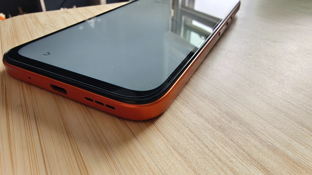 Close-up of a smartphone with an orange case, resting on a wooden surface, showing the bottom edge with ports.