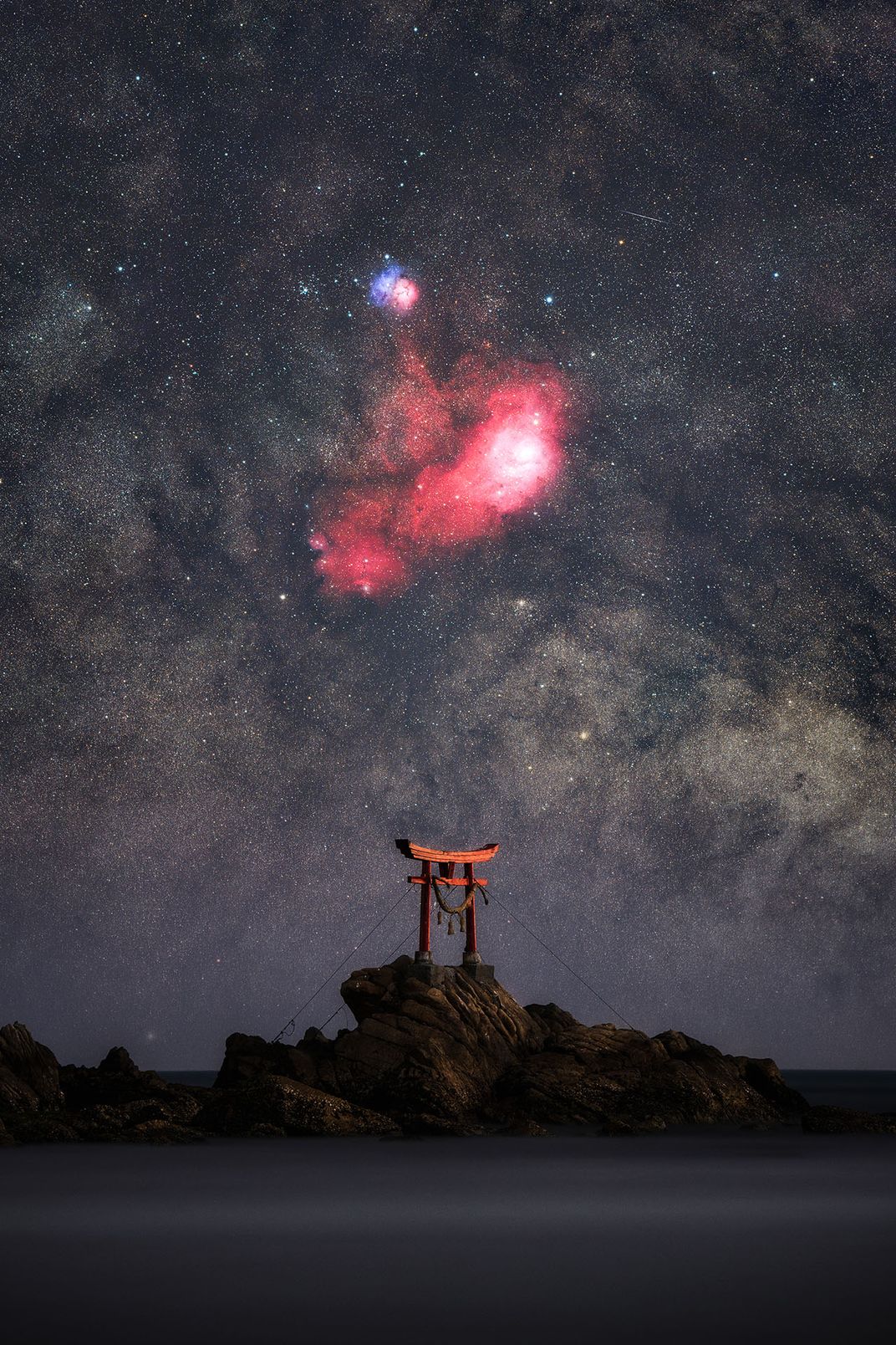 Torii gate sits on rocks in the ocean underneath stars and glowing red and blue patches