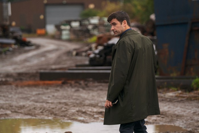 A person in a long dark green coat walks through a muddy industrial yard, glancing back over their shoulder while holding a notebook or folder at their side. Puddles cover the ground, with stacks of metal and industrial equipment blurred in the background, creating a damp, overcast setting with a tense, investigative mood.
