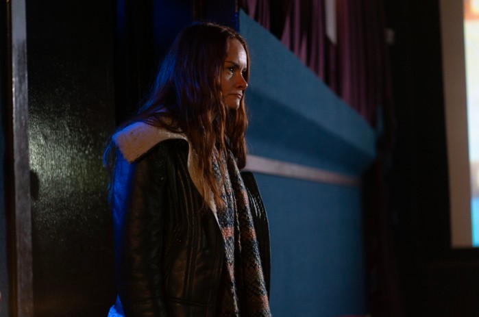 A person with long auburn hair stands inside a dimly lit venue, wearing a black shearling-lined jacket and a multicoloured knitted scarf. They look off to one side with a tense, thoughtful expression, while dark curtains and the edge of a projection screen in the background suggest a cinema, theatre or community hall.
