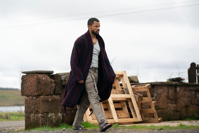 A person wearing a grey vest, beige cargo trousers and sandals walks outdoors wrapped in a dark striped dressing gown. They pass a low stone wall with stacked wooden pallets beside it, under an overcast sky, with open countryside visible in the background. Their serious expression and casual clothing create a sense of urgency or unease.