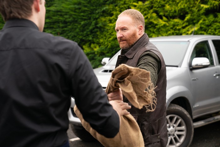 A red-bearded person wearing a dark jacket and brown work vest hands a folded hessian sack to another person whose back is to the camera. They stand in a car park beside a silver pickup truck, with tall green hedges in the background, suggesting a practical exchange taking place outdoors.