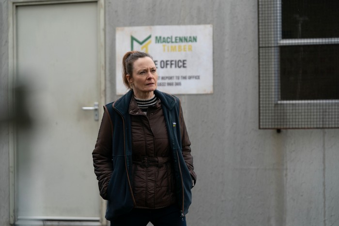 A person wearing a brown quilted jacket, striped high-neck top and dark sleeveless fleece stands outside a portable site office with their hands in their pockets. A sign for "MacLennan Timber" is mounted on the wall behind them, and their focused expression suggests they are watching or waiting in an industrial worksite setting.