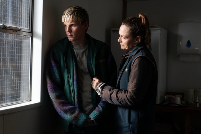A young blond person stands by a frosted window inside a modest office or staff room, looking outside with a worried expression. Beside them, an older person wearing a dark fleece over a brown jacket gently holds the front of their cardigan in a reassuring gesture. The subdued lighting and sparse surroundings create a quiet, intimate moment of comfort and concern.