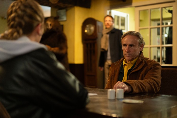 Inside a warmly lit pub, a grey-haired person in a brown jacket sits at a wooden table facing another person whose back is to the camera. Their expression is serious and focused, while another figure stands in the open doorway behind them. A grandfather clock and a large taxidermy bear in the background add to the distinctive, slightly unsettling atmosphere.