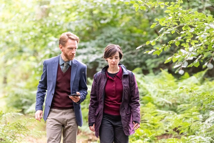 Two people walk side by side along a woodland path surrounded by lush green trees and ferns. One wears a blue blazer and burgundy jumper while looking at a smartphone, and the other wears a dark purple jacket over a burgundy top, looking thoughtfully ahead. Their serious expressions and quiet conversation suggest they are discussing something important as they move through the forest.