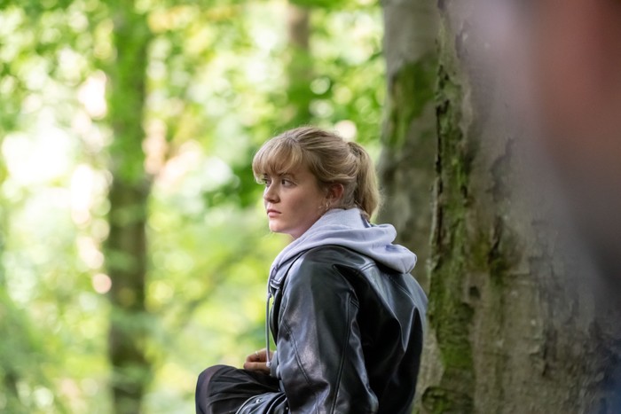 A young person with blonde hair tied back sits against a tree in a sunlit woodland, wearing a black leather jacket over a light grey hooded sweatshirt. They look off into the distance with a pensive, slightly anxious expression, while the softly blurred green forest around them creates a quiet, reflective atmosphere.