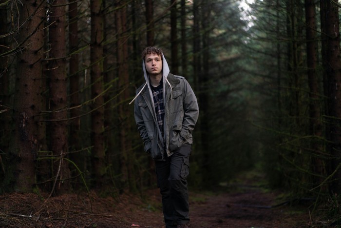 A young person with curly hair walks alone along a narrow path through a dense conifer forest, wearing a grey jacket layered over a hooded sweatshirt and checked shirt. With their hands in their pockets and a serious expression, they move beneath the tall trees, while the dim light and shadowy woodland create a quiet, isolated atmosphere.