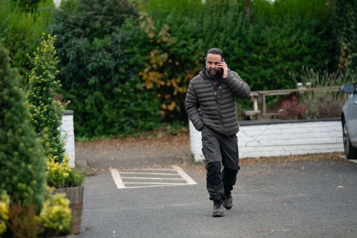 A bearded person wearing a grey quilted jacket and dark work trousers walks briskly across a car park while talking on a mobile phone. One hand is in their pocket, and trees, shrubs and a low white wall frame the background, creating the feel of a quiet residential or business property.
