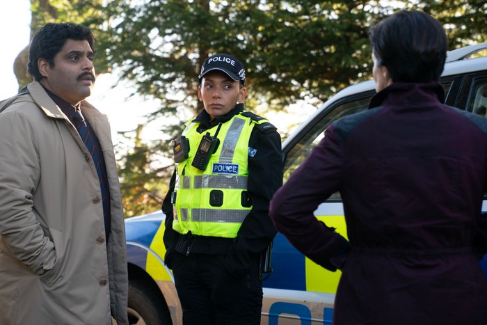Laura Donnelly as DI Monica Kennedy stands beside a marked police car, speaking with a uniformed police officer and another man in a wooded area in The Dark.
