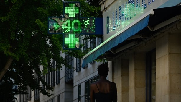 A woman walks by an illuminated green cross of a pharmacy reading 