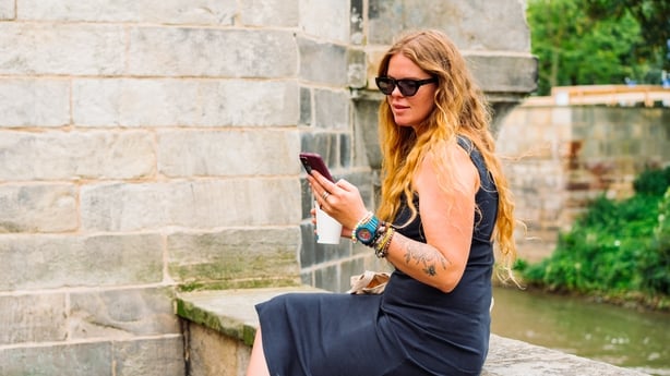Modern woman with long hair and boho accessories sitting outdoors by the river, using a smartphone while holding a takeaway coffee. Technology and lifestyle concept in a relaxed natural setting. Embracing digital life, freedom, and connectivity on the go.