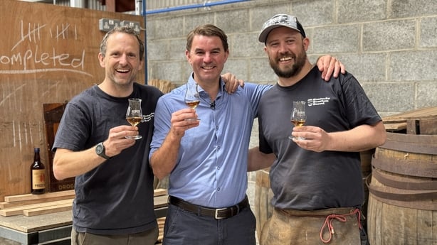 Three smiling men hold glasses of whiskey in a cooperage