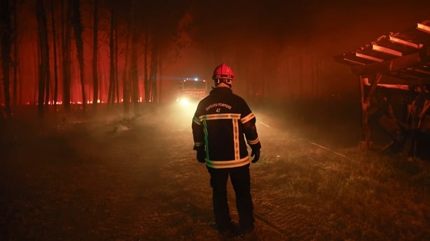A lone firefighter watches a fire truck drive towards him as a wildfire glows among trees in the background