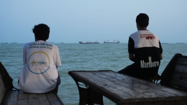 Two youths sit on the shoreline as ships are seen anchored in the Strait of Hormuz on August 10, 2026 off the coast of Bandar Abbas, Iran