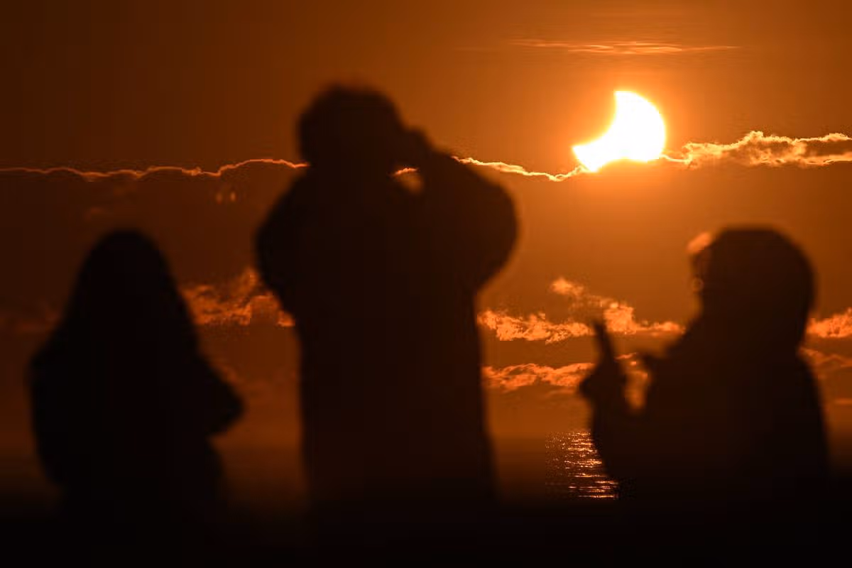Silhouettes of individuals standing in a dimly lit outdoor setting at night, with a fire burning in the background. One individual is engaged in a conversation on a mobile device.