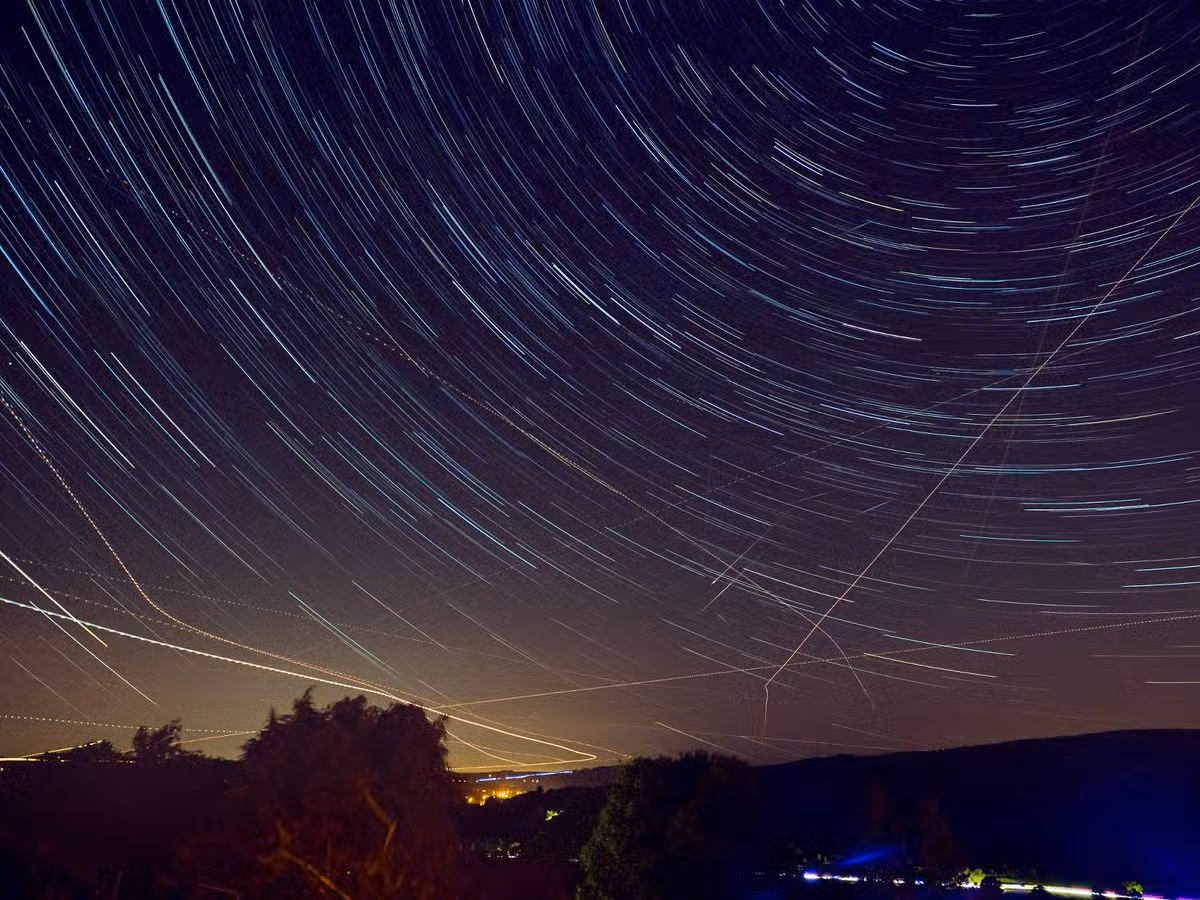 A nighttime landscape featuring a star-studded sky with prominent star trails, indicative of long-exposure photography. The silhouettes of terrestrial features are visible against the horizon, bathed in the soft glow of twilight.