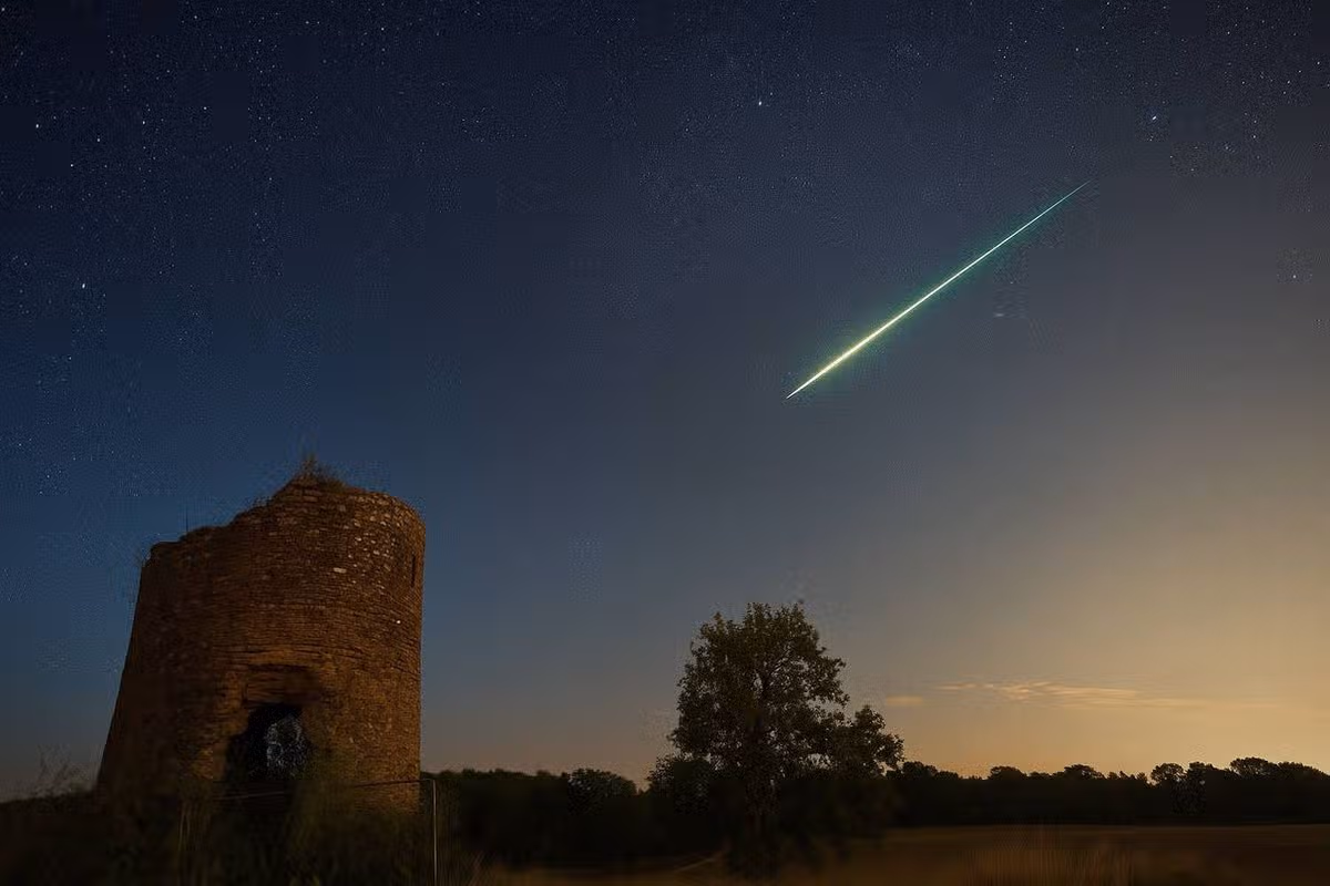 An image depicting a bright meteor streaking across a twilight sky, with a historic structure and a tree in the foreground.