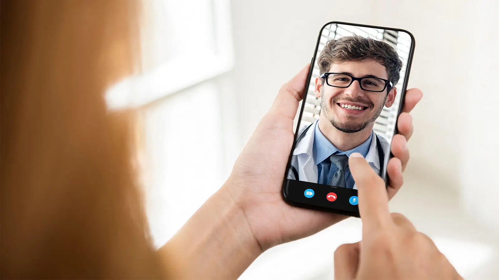 A photo of a woman holding a smartphone displaying a male physician.