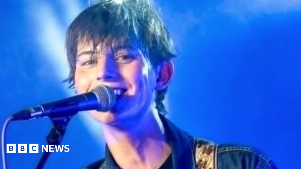 A close-up image of Kit Bottomley with short brown hair, who is smiling and singing into a microphone against a blue backdrop.