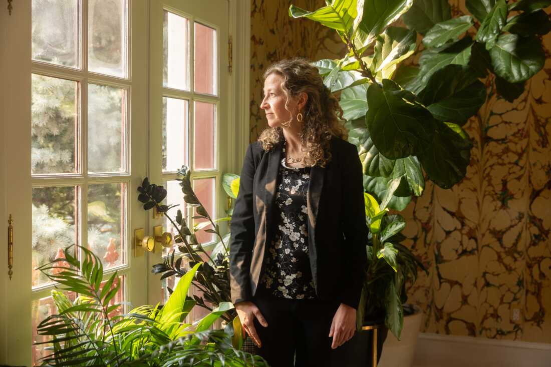 Sudbury looks out of a glass paneled door while standing in front of large potted plants.