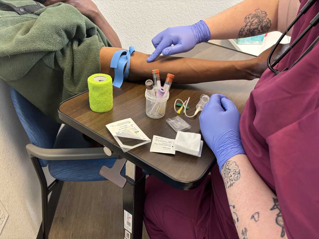 The mid-range photograph focuses on the arm and hands of medical assistant Izzy Kadrmas and patient Tywon Pugh.