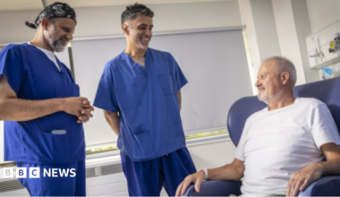 Two men dressed in blue scrubs are on the left and in the middle and standing up. They are looking towards Hall, who has a beard and grey hair and is sitting on a blue chair on the right.