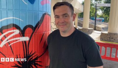 A man with short brown hair and a black T-shirt is smiling while standing next to a pillar painted blue with a red poppy design.
