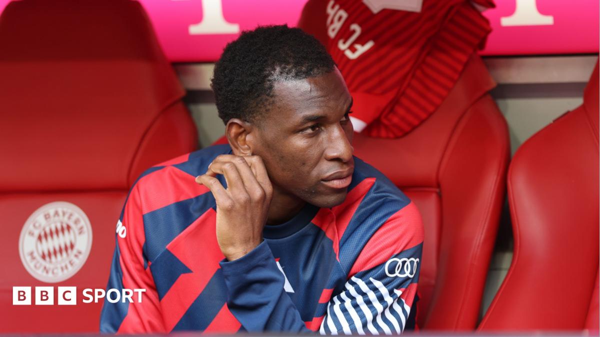 Nicolas Jackson, wearing a blue and red training top, sits on the Bayern Munich bench before a Bundesliga match against Cologne