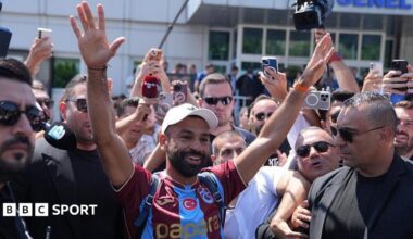 Mohamed Salah smiling with his hands in the air in a Trabzonspor shirt while being surrounded by people outside the airport