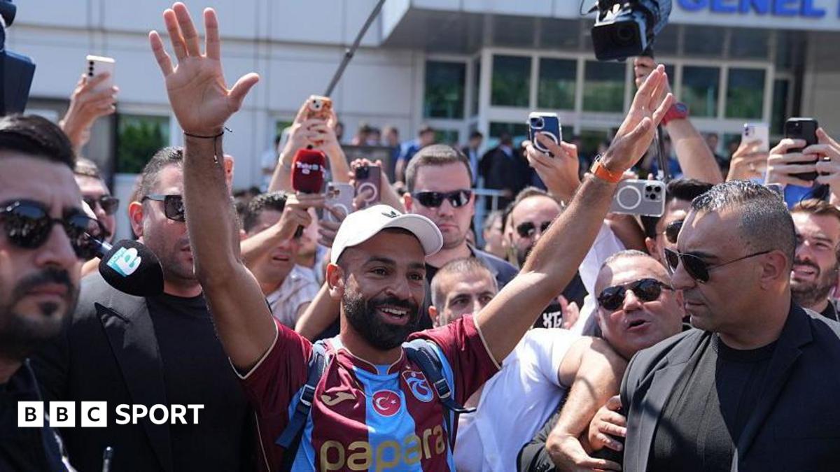 Mohamed Salah smiling with his hands in the air in a Trabzonspor shirt while being surrounded by people outside the airport