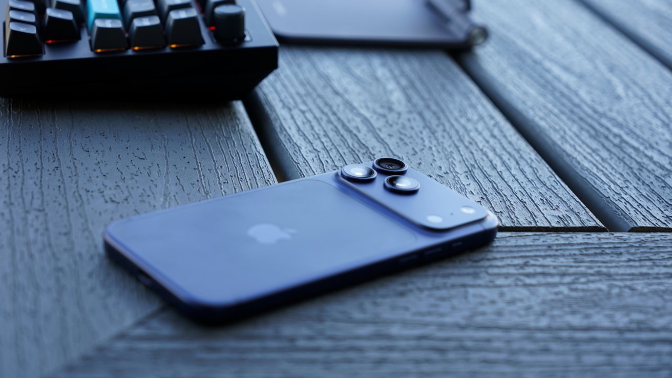 Blue smartphone lying face down on a textured gray wooden surface, camera lenses visible, with a keyboard and another device blurred in the background