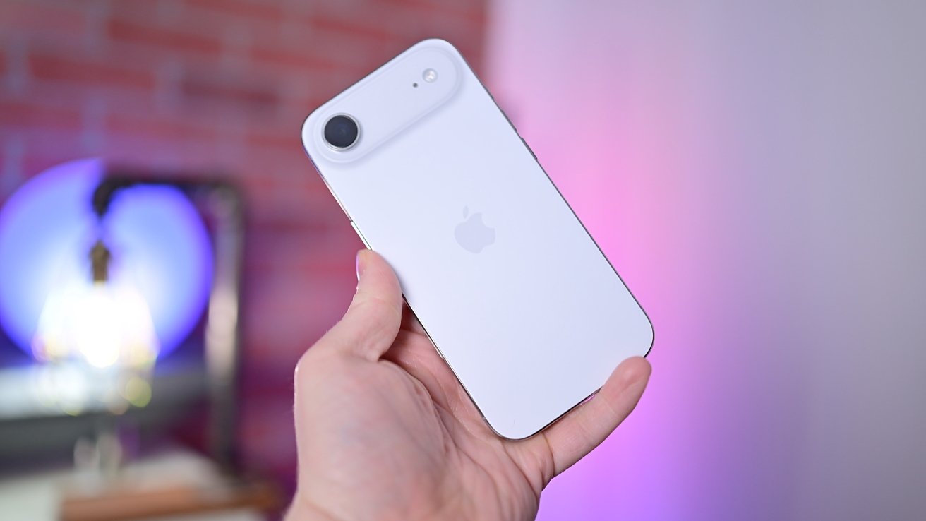 Hand holding a white iPhone with a single rear camera against a softly blurred background featuring pink and purple lighting and a partially visible brick wall
