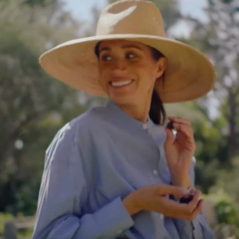 Smiling woman wearing a wide-brimmed hat and blue shirt, holding berries outdoors in a garden setting.