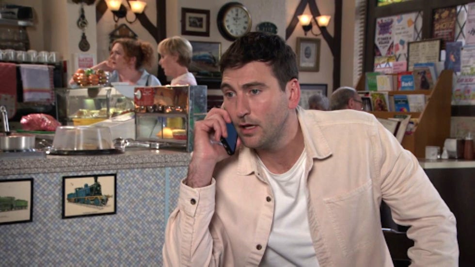 coronation street kit green man talking on phone at a busy café counter.