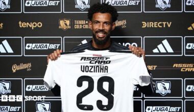 Vozinha, smiling and holding a white Colo Colo shirt with Vozihna 29 on the back in front of a black board of sponsor logos