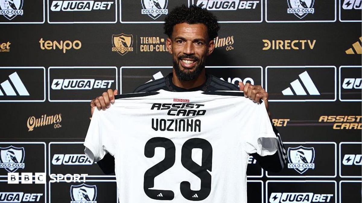 Vozinha, smiling and holding a white Colo Colo shirt with Vozihna 29 on the back in front of a black board of sponsor logos