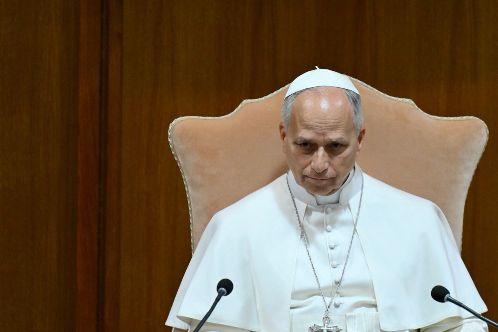 Pope Leo white in a robe, skullcap, and cross necklace, seated looking downward in a high-backed chair between two microphones against a wooden wall background.