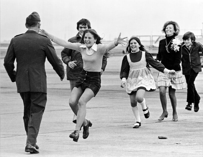 A man in military uniform walks toward a joyful girl running with arms outstretched, followed by smiling family members, on an airport tarmac.