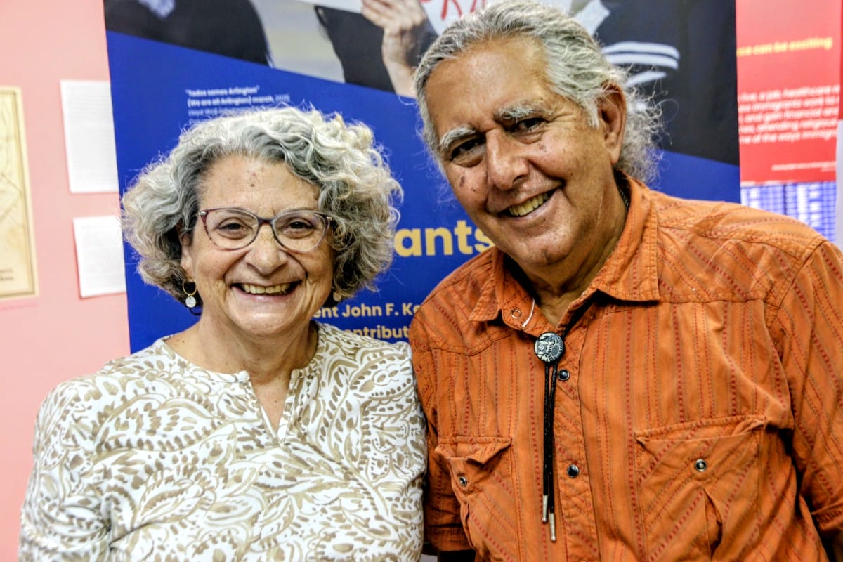 Smiling elderly couple posing together indoors, woman with glasses and patterned blouse, man in orange shirt with bolo tie.