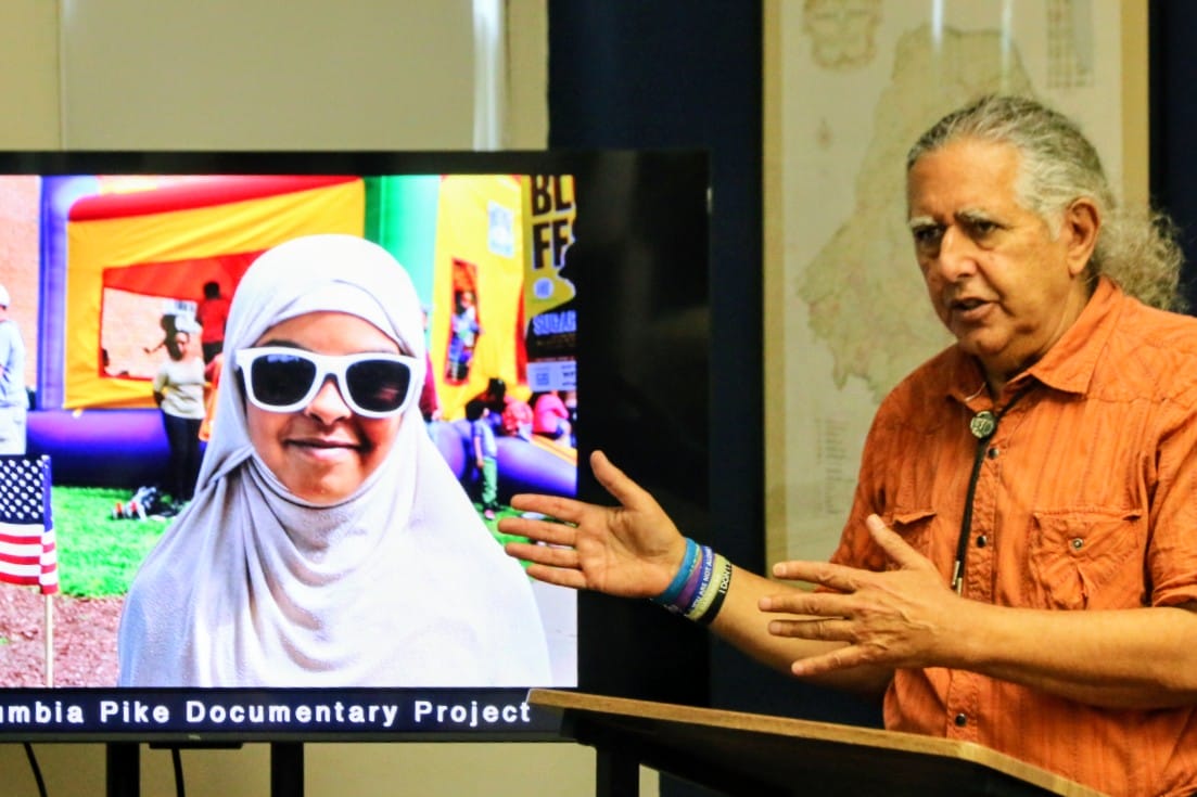 Presenter in an orange shirt gestures toward a TV screen showing a smiling girl in a hijab and white sunglasses, part of a documentary project display.