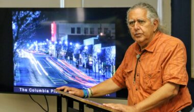 Man in an orange shirt stands at a lectern beside a large TV displaying a nighttime city street scene.