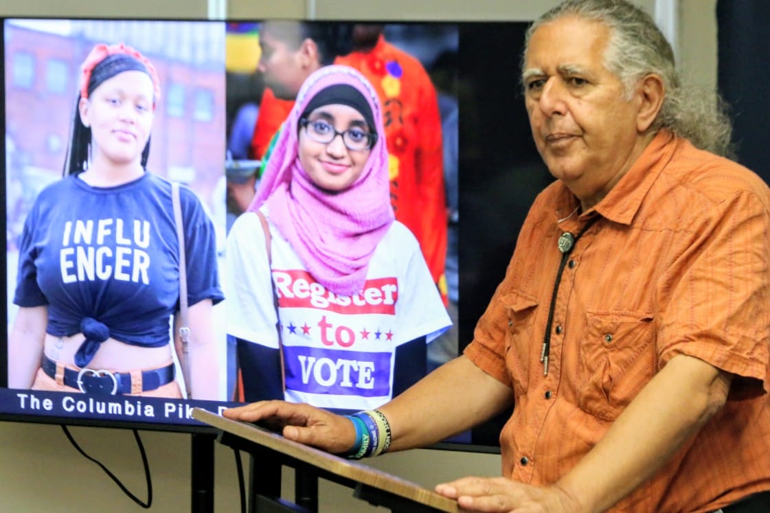 Older man in orange shirt speaks at a podium beside a large screen showing two young women wearing campaign-style shirts promoting voting, including hijab-wearing student on the right and a shirt reading 'Influencer' on the left.