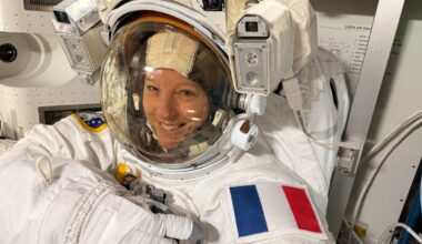 A woman wearing an ESA pressurised space suit smiles and points to her left shoulder, on which a patch of the French flag is displayed. She is Sophie Adenot.