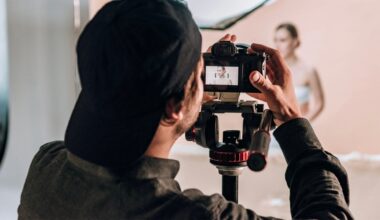 A photographer wearing a black baseball cap looks through the viewfinder of a camera mounted on a tripod, focusing on a model in the background.