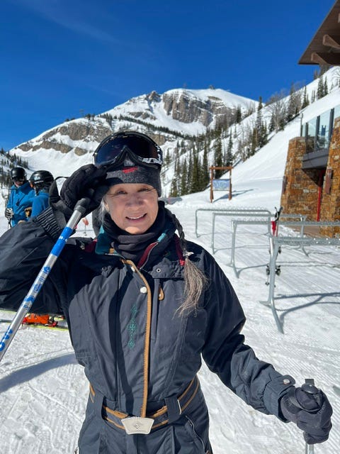 Smiling skier in winter gear on a slope with mountains in the background, holding a ski pole and looking towards the camera. Smiling skier in winter gear on a slope with mountains in the background, holding a ski pole and looking towards the camera.