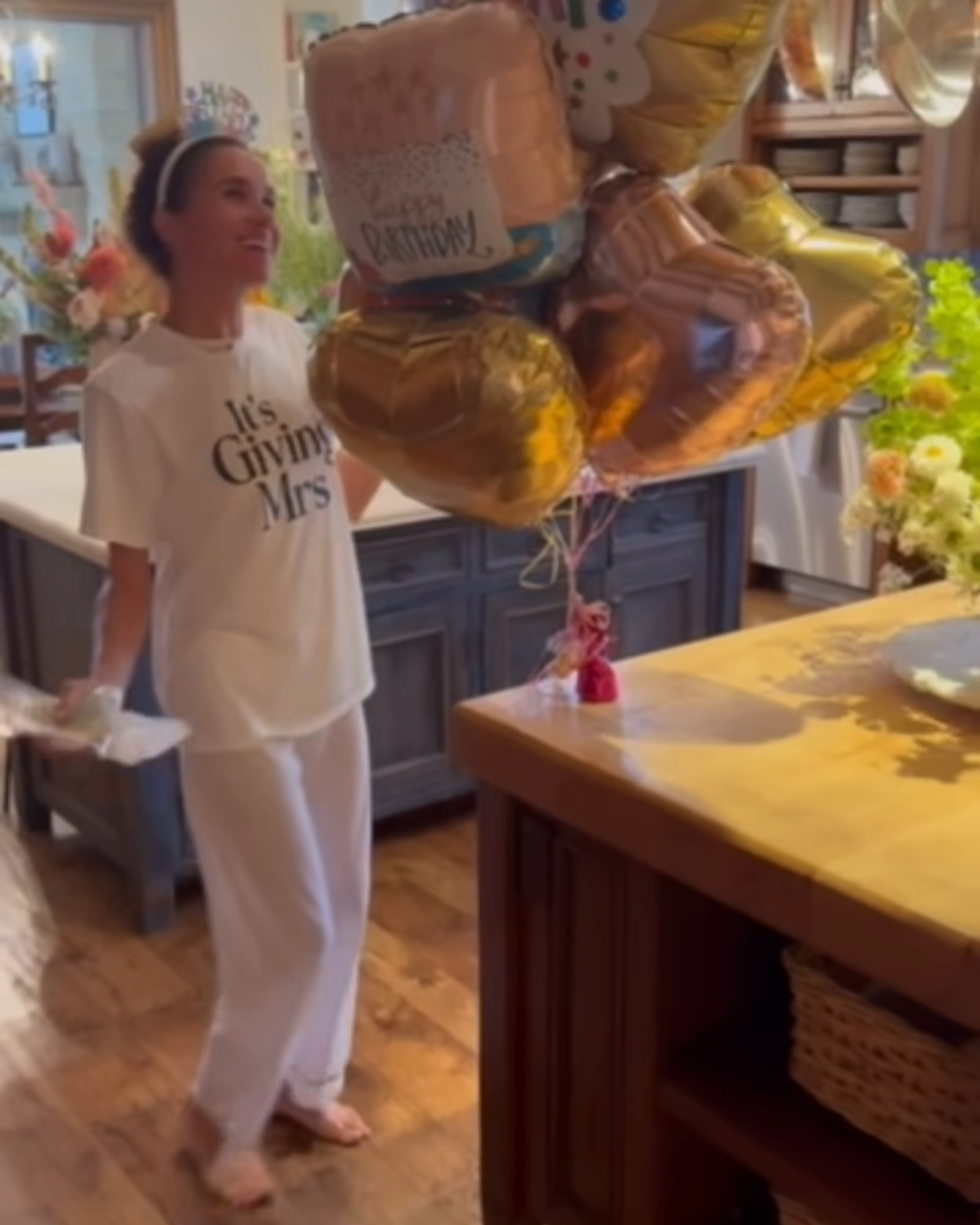 Person in a white outfit joyfully holding a bundle of colorful birthday balloons in a cheerful indoor setting.