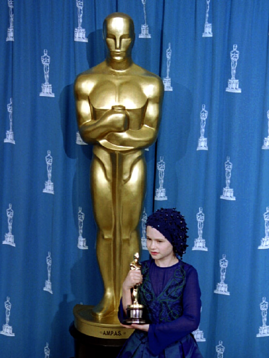 A little girl holds a gold Oscar, dwarfed by a replica of the statue behind her.